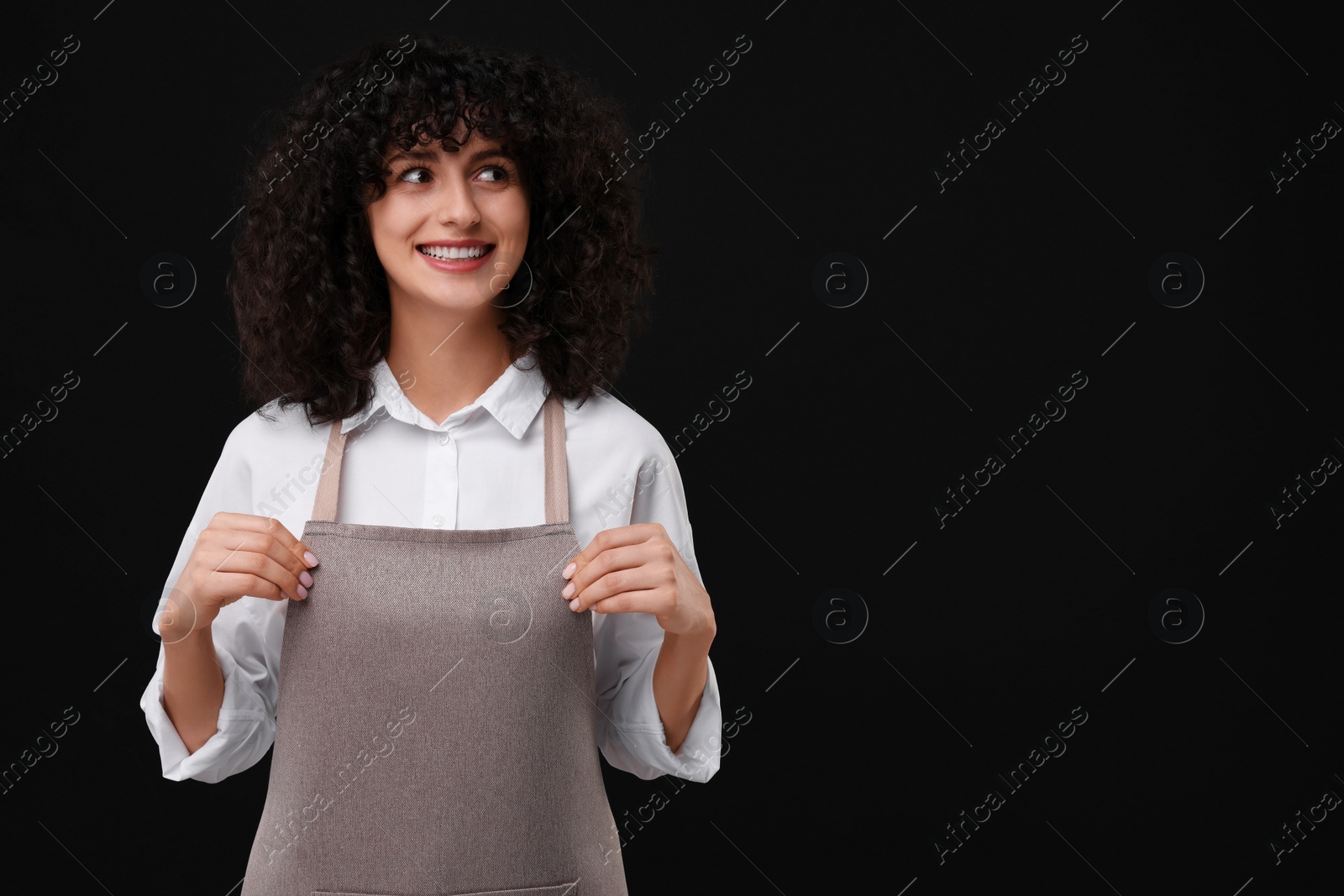 Photo of Happy woman wearing kitchen apron on black background, space for text. Mockup for design