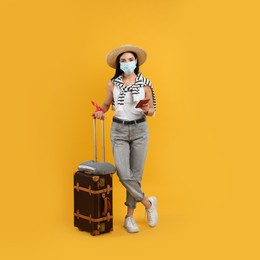 Photo of Female tourist in medical mask with suitcase, ticket and passport on yellow background. Travelling during coronavirus pandemic