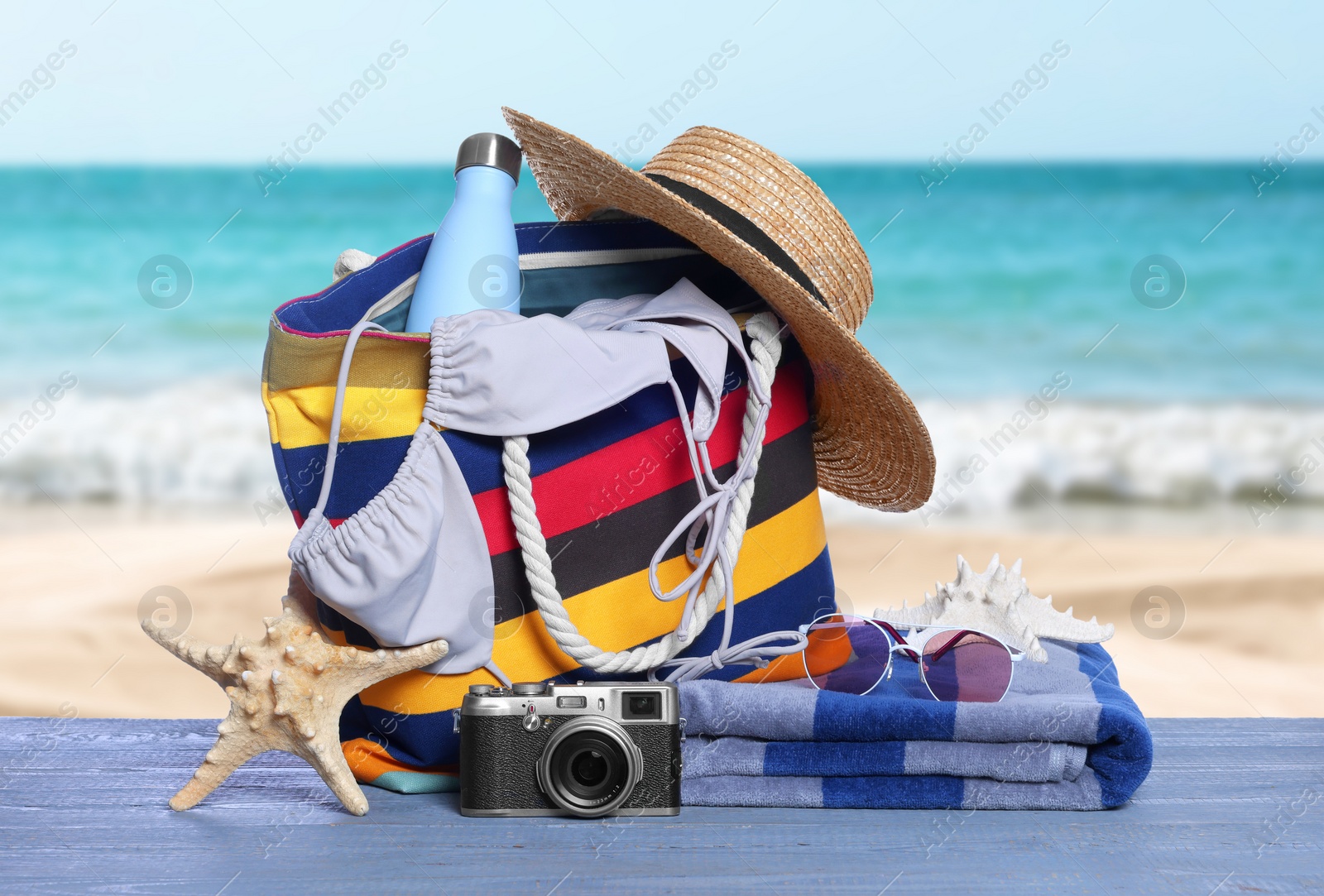 Image of Stylish striped bag and other beach accessories on blue wooden table near sea