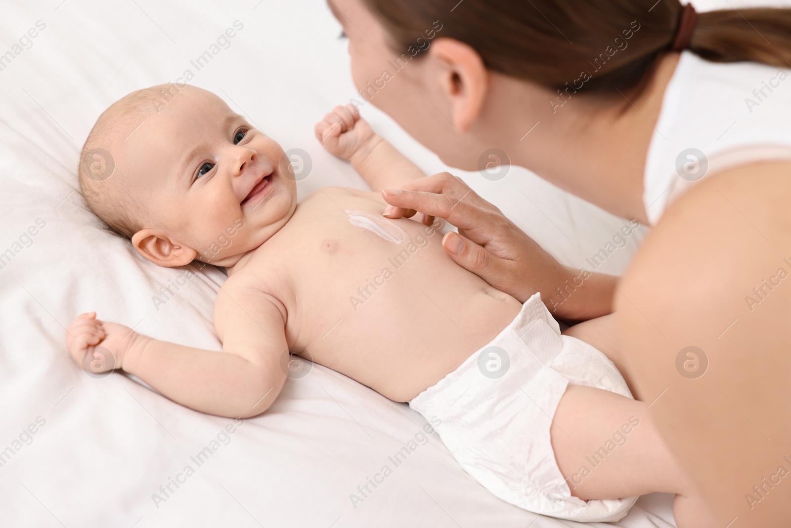 Photo of Woman applying body cream onto baby`s skin on bed, closeup