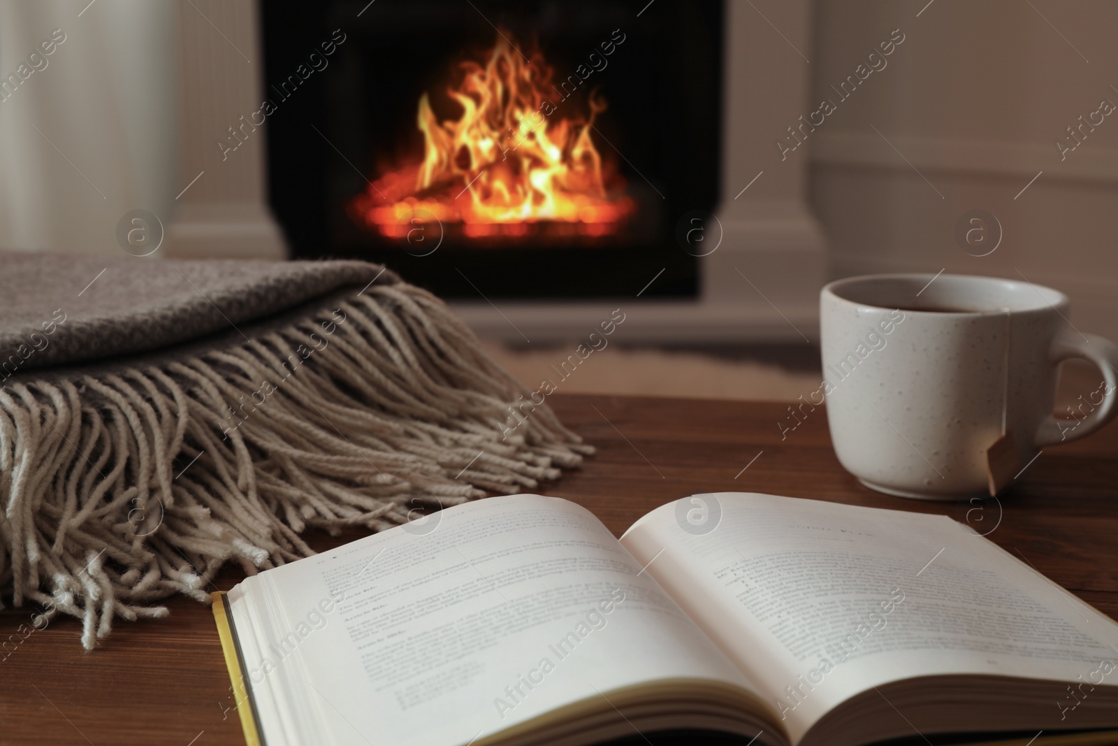 Photo of Cup of hot tea and book on wooden table near fireplace at home. Cozy atmosphere