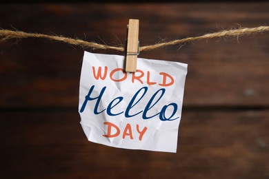 Image of Crumpled paper note with phrase World Hello Day hanging on twine against wooden background