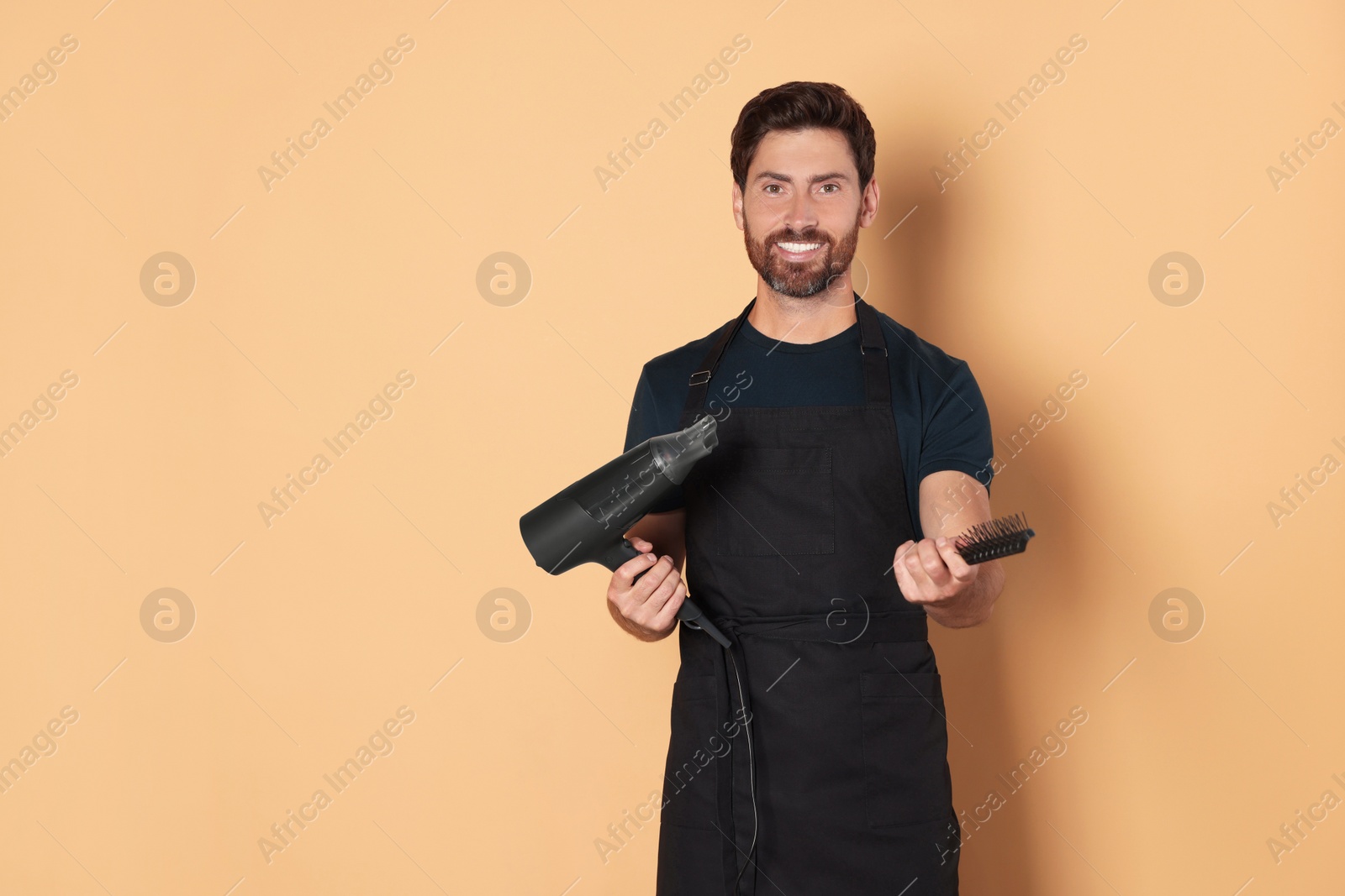 Photo of Smiling hairdresser in apron holding dryer and brush on light brown background, space for text