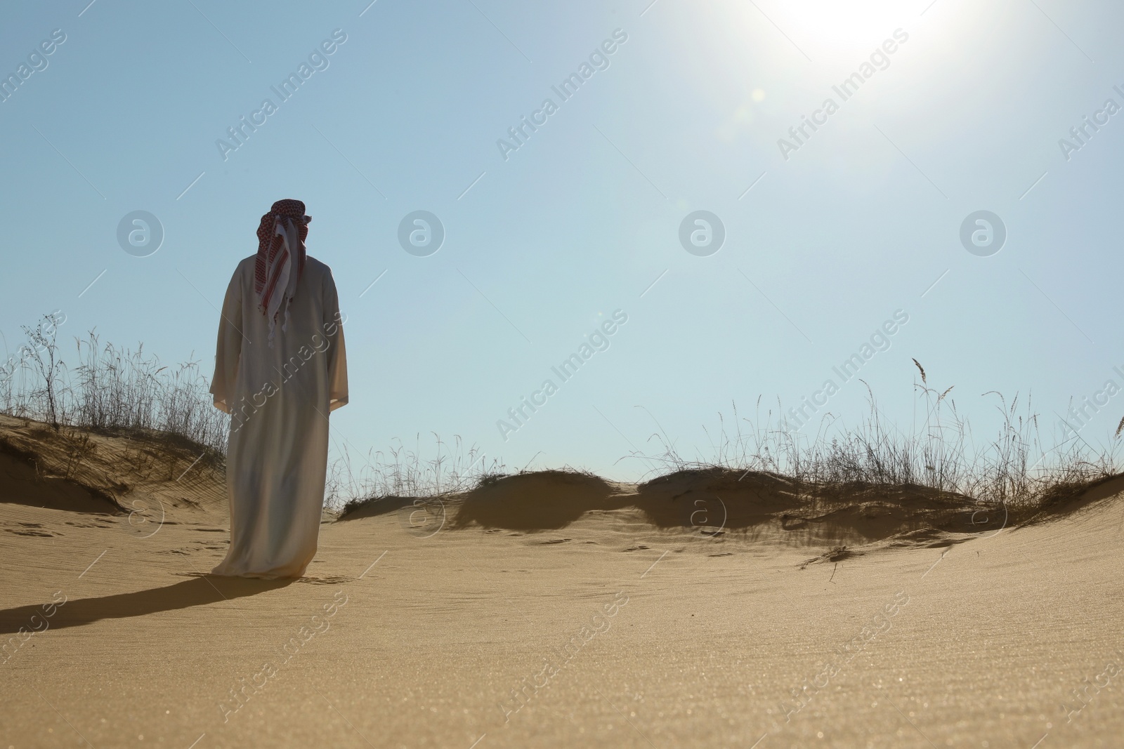 Photo of Man in arabic clothes walking through desert on sunny day, back view