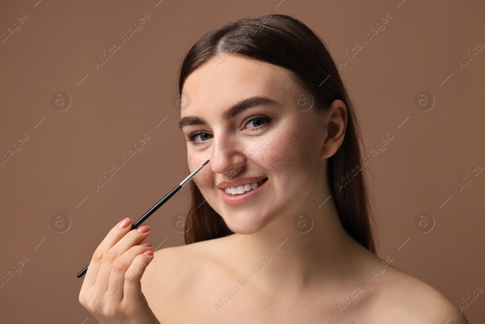 Photo of Smiling woman drawing freckles with brush on brown background