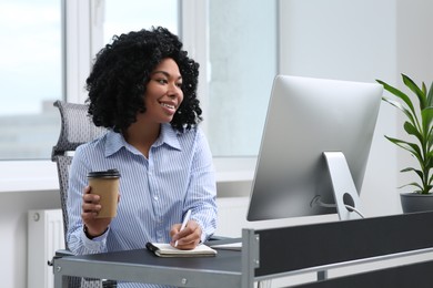 Young woman with cup of drink taking notes near computer at table in office