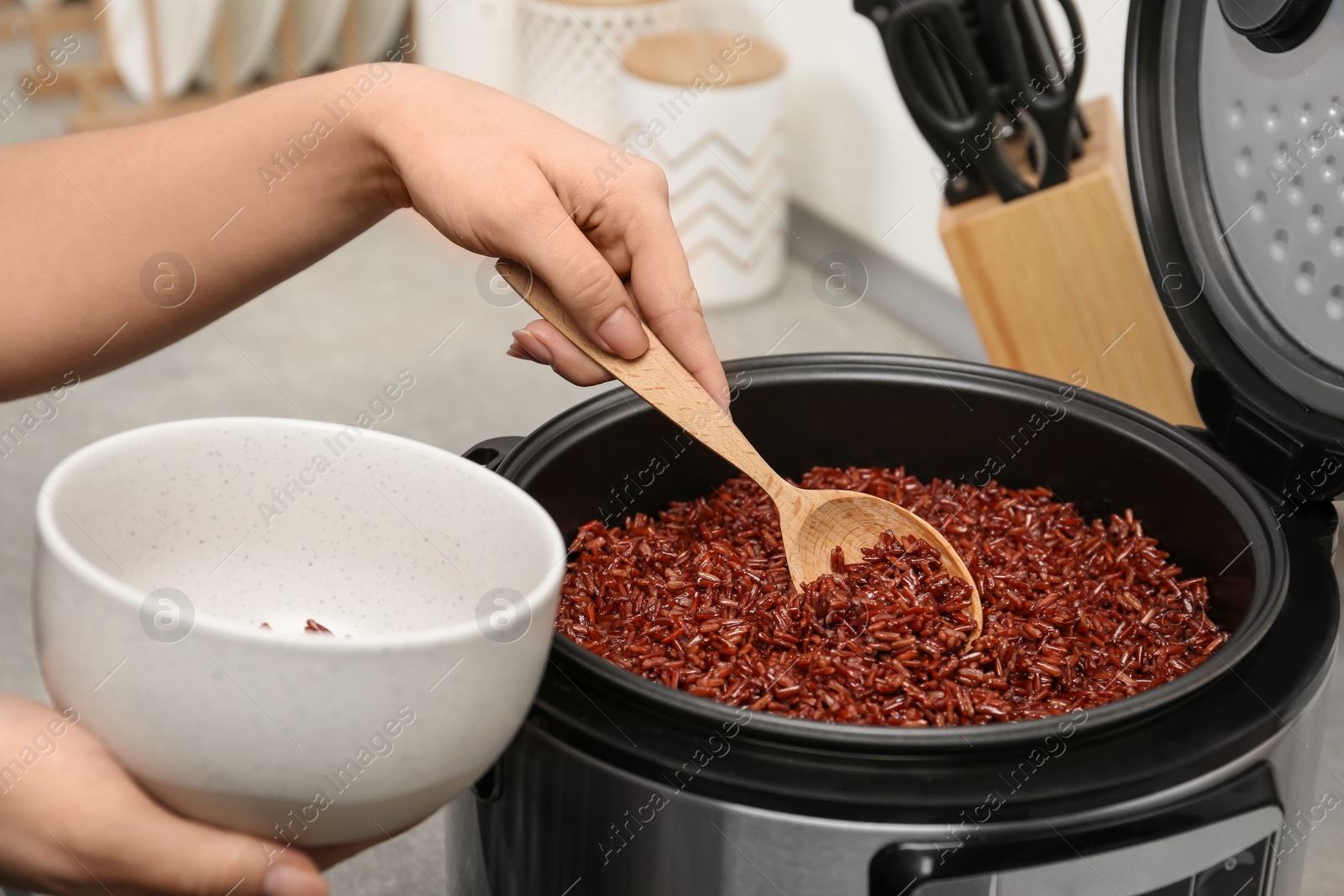 Photo of Woman putting brown rice into bowl from multi cooker in kitchen, closeup