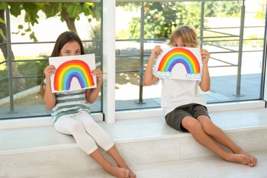 Photo of Little children holding rainbow paintings near window indoors. Stay at home concept