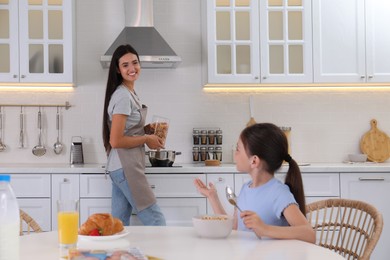 Little girl having breakfast while her mother cooking food in kitchen. Single parenting