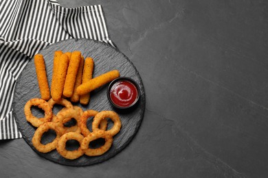 Photo of Tasty ketchup, onion rings and cheese sticks on dark textured table, top view. Space for text