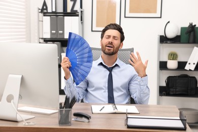 Bearded businessman waving blue hand fan to cool himself at table in office