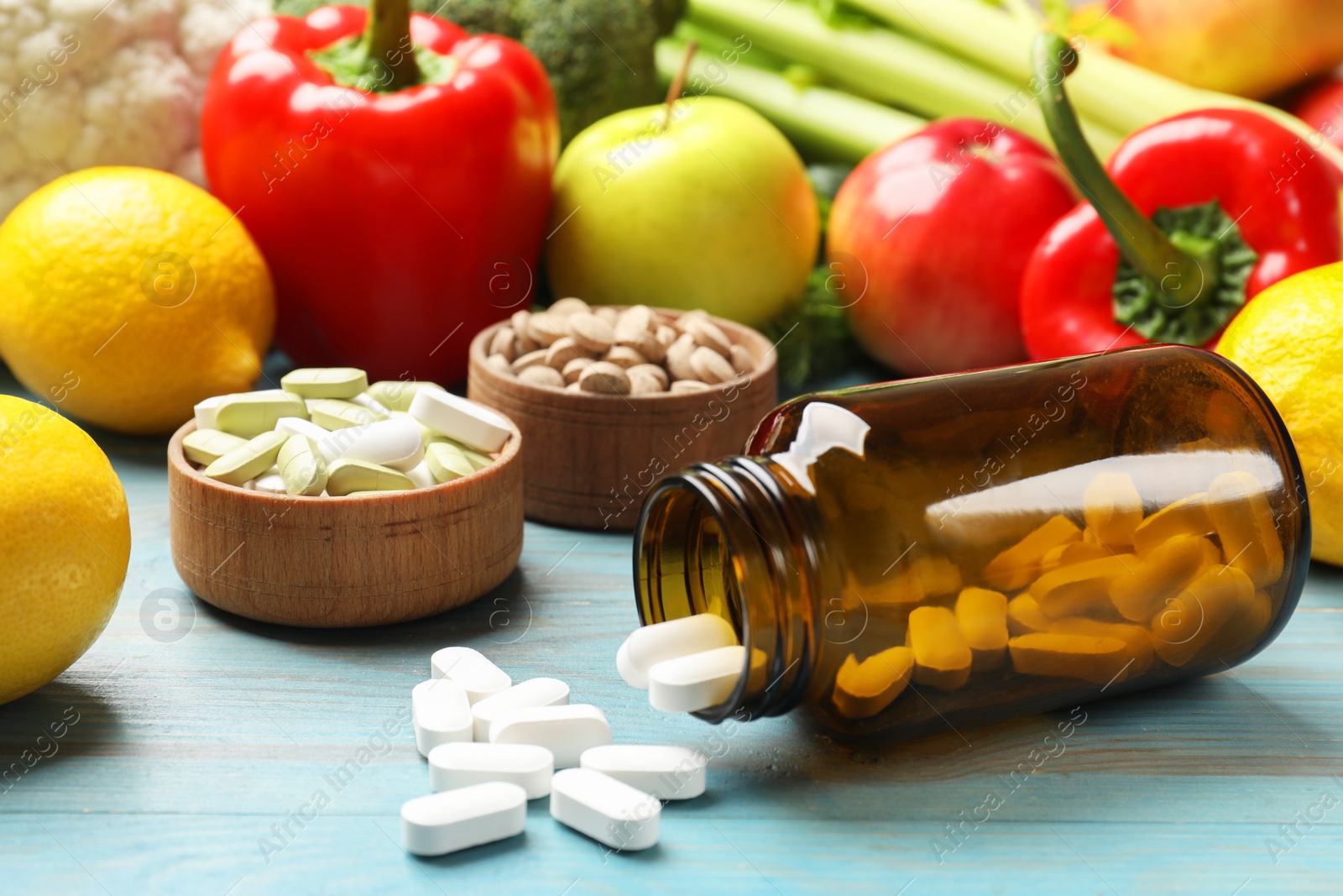 Photo of Dietary supplements. Overturned bottle and bowls with different pills near food products on light blue wooden table