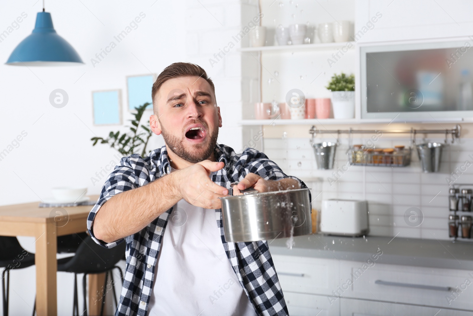 Photo of Emotional young man holding saucepan under water leakage from ceiling in kitchen, space for text. Plumber service