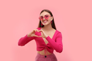 Photo of Pink look. Beautiful woman in sunglasses making heart with hands on color background
