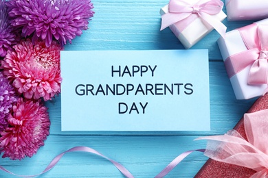 Photo of Beautiful flowers, gift boxes and card with phrase Happy Grandparents Day on light blue wooden background, flat lay