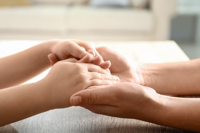 Photo of Mother holding hands with her child at wooden table indoors, closeup. Happy family