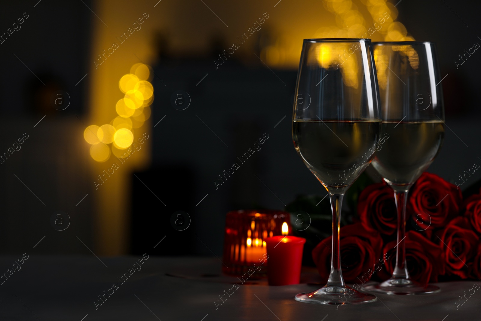 Photo of Glasses of white wine, burning candles and rose flowers on grey table against blurred lights, space for text. Romantic atmosphere