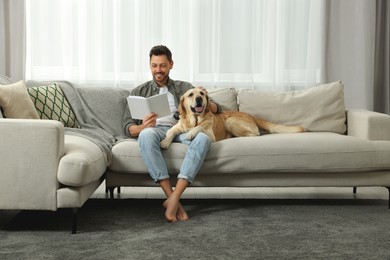 Man reading book on sofa near his cute Labrador Retriever at home. Space for text