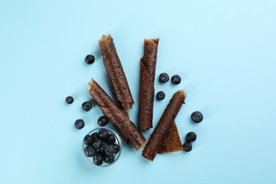 Delicious fruit leather rolls and blueberries on light blue background, flat lay