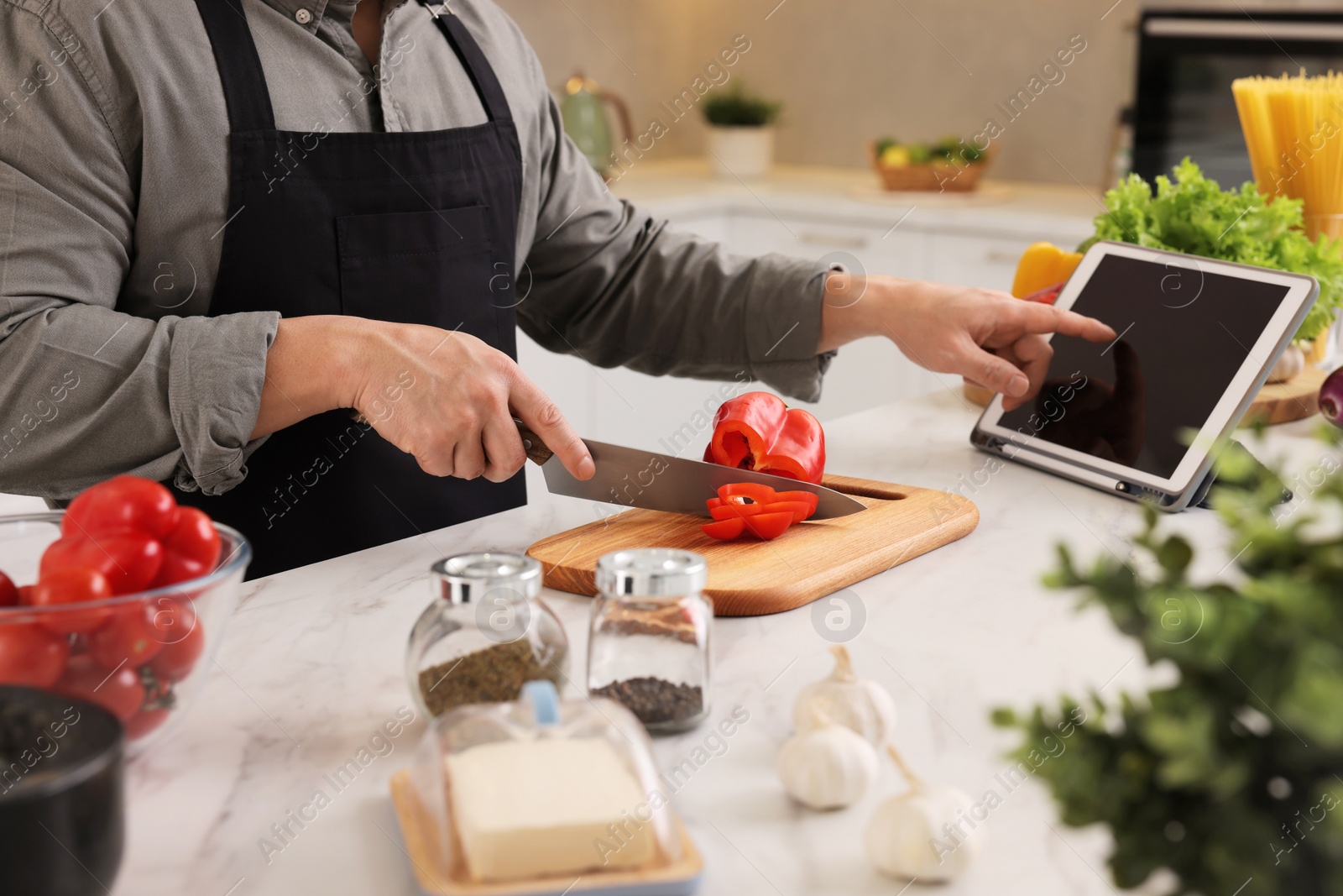 Photo of Cooking process. Man using tablet while cutting fresh bell pepper at white marble countertop in kitchen, closeup