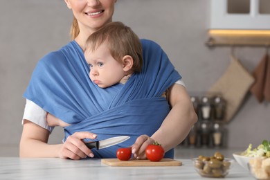 Mother cutting tomatoes while holding her child in sling (baby carrier) indoors