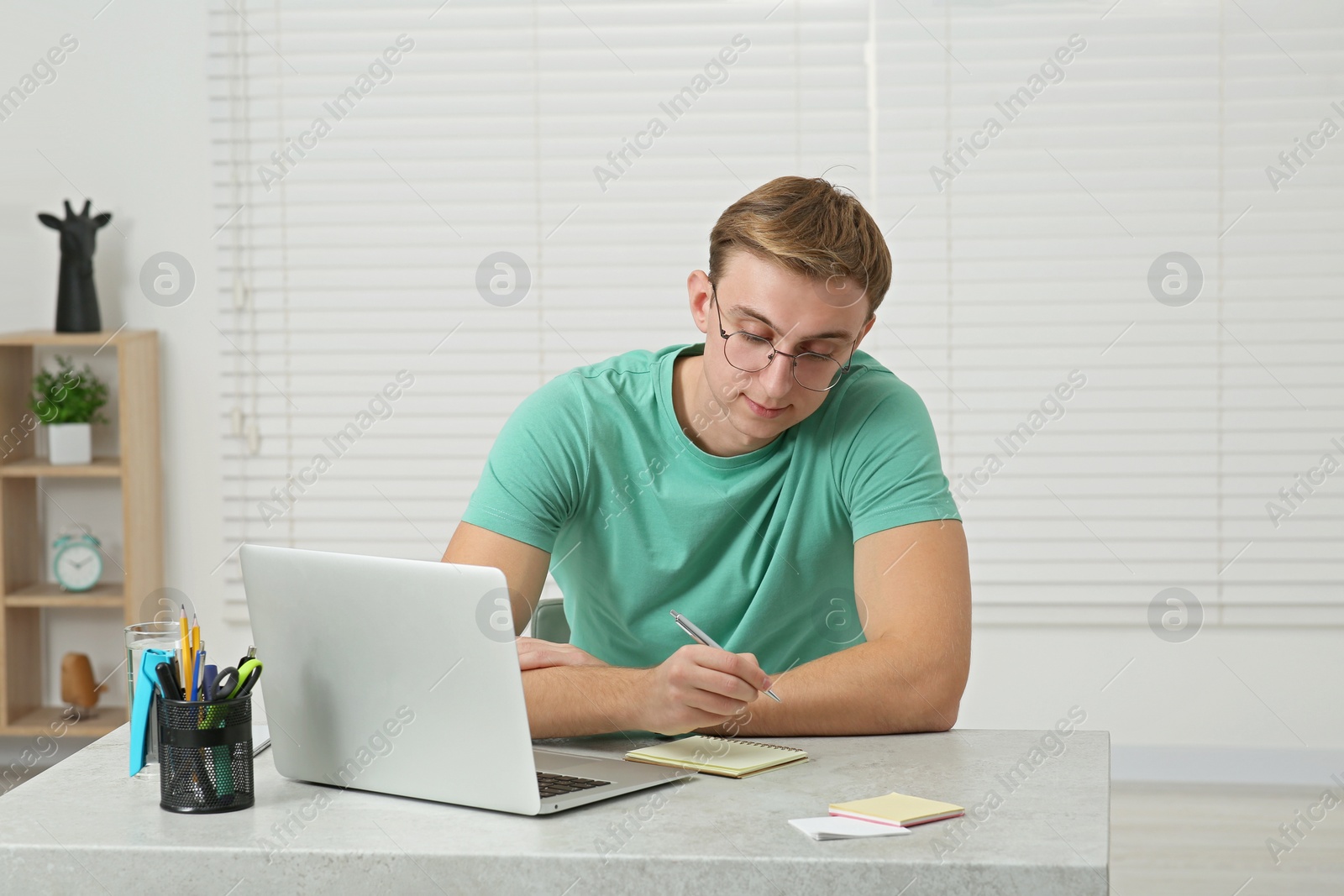 Photo of Young man with laptop writing in notebook at table indoors