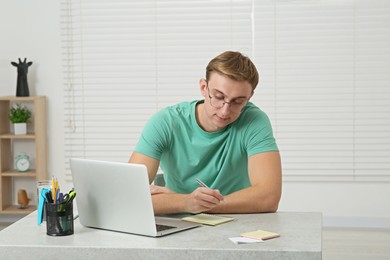 Photo of Young man with laptop writing in notebook at table indoors