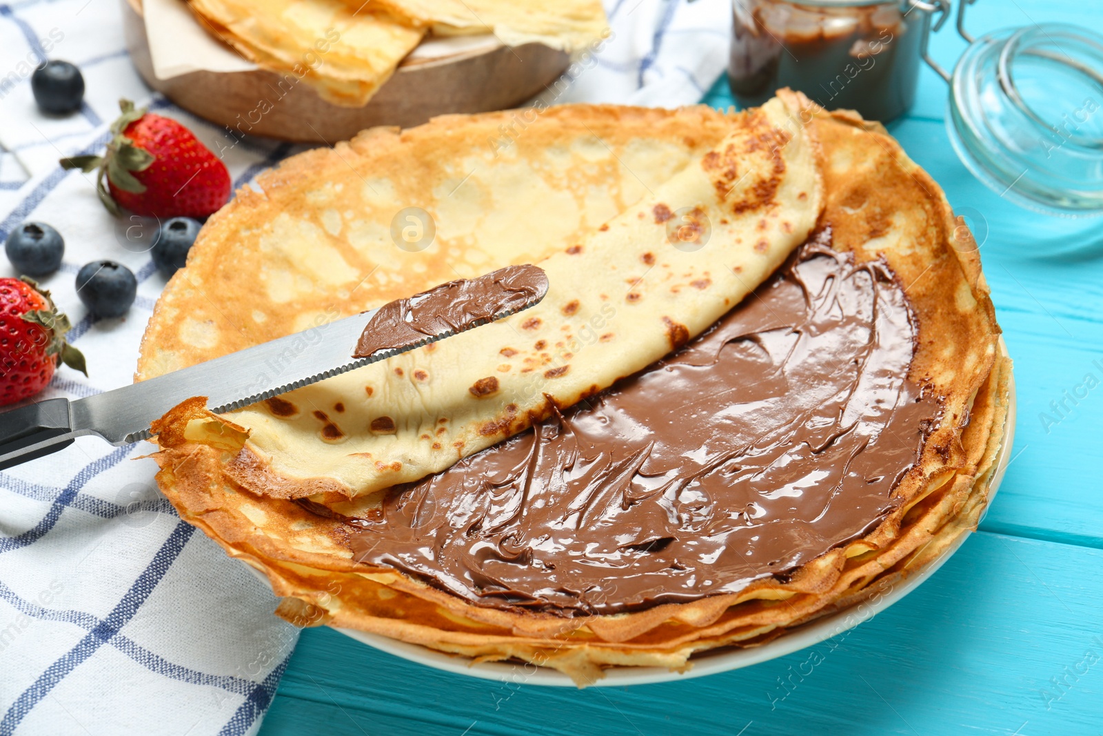 Photo of Tasty crepes with chocolate paste and berries served on turquoise wooden table, closeup