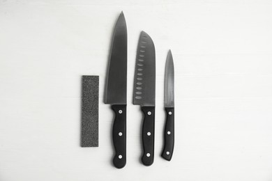 Photo of Sharpening stone and set of knives on white wooden table, flat lay