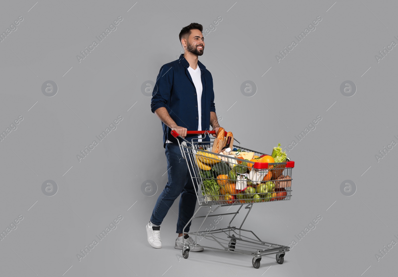 Photo of Happy man with shopping cart full of groceries on light grey background