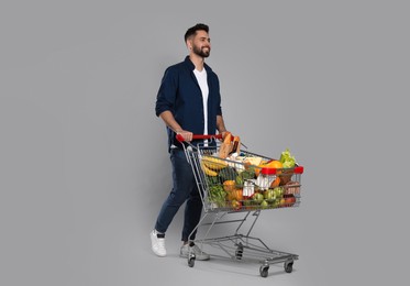 Photo of Happy man with shopping cart full of groceries on light grey background