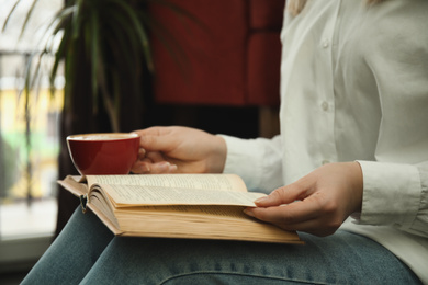 Woman with cup of coffee reading book indoors, closeup