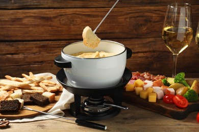 Dipping piece of bread into fondue pot with melted cheese at wooden table with snacks, closeup