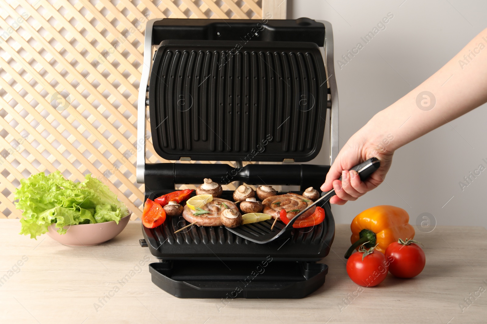 Photo of Woman cooking homemade sausages with bell peppers and mushrooms on electric grill at wooden table, closeup