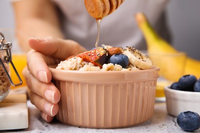 Woman adding honey to oatmeal with blueberries, almonds, banana and fig pieces at white table, closeup