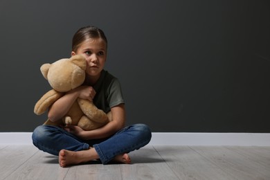 Photo of Child abuse. Upset girl with toy sitting on floor near grey wall, space for text