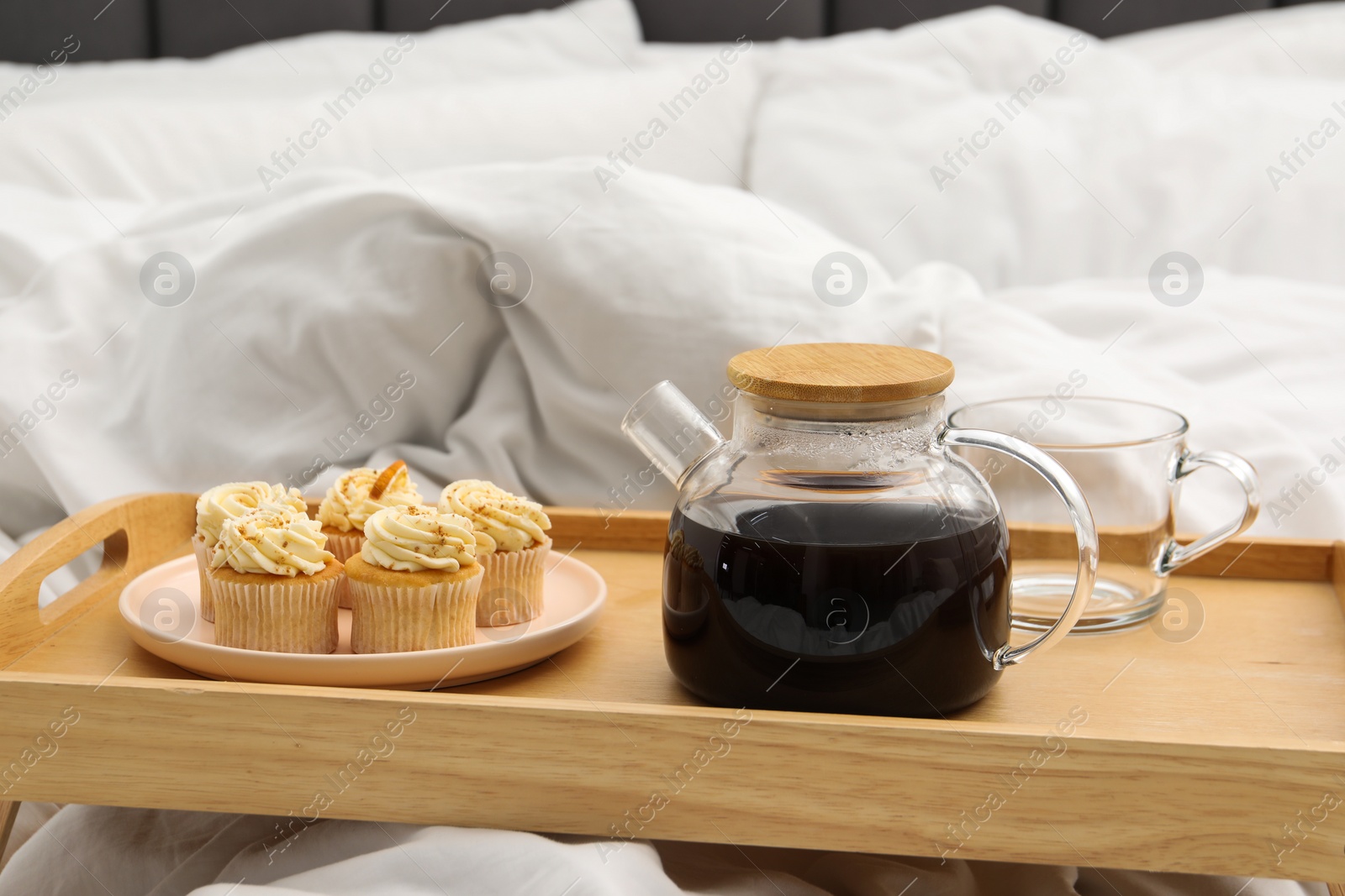 Photo of Wooden tray with aromatic tea and tasty cupcakes on bed