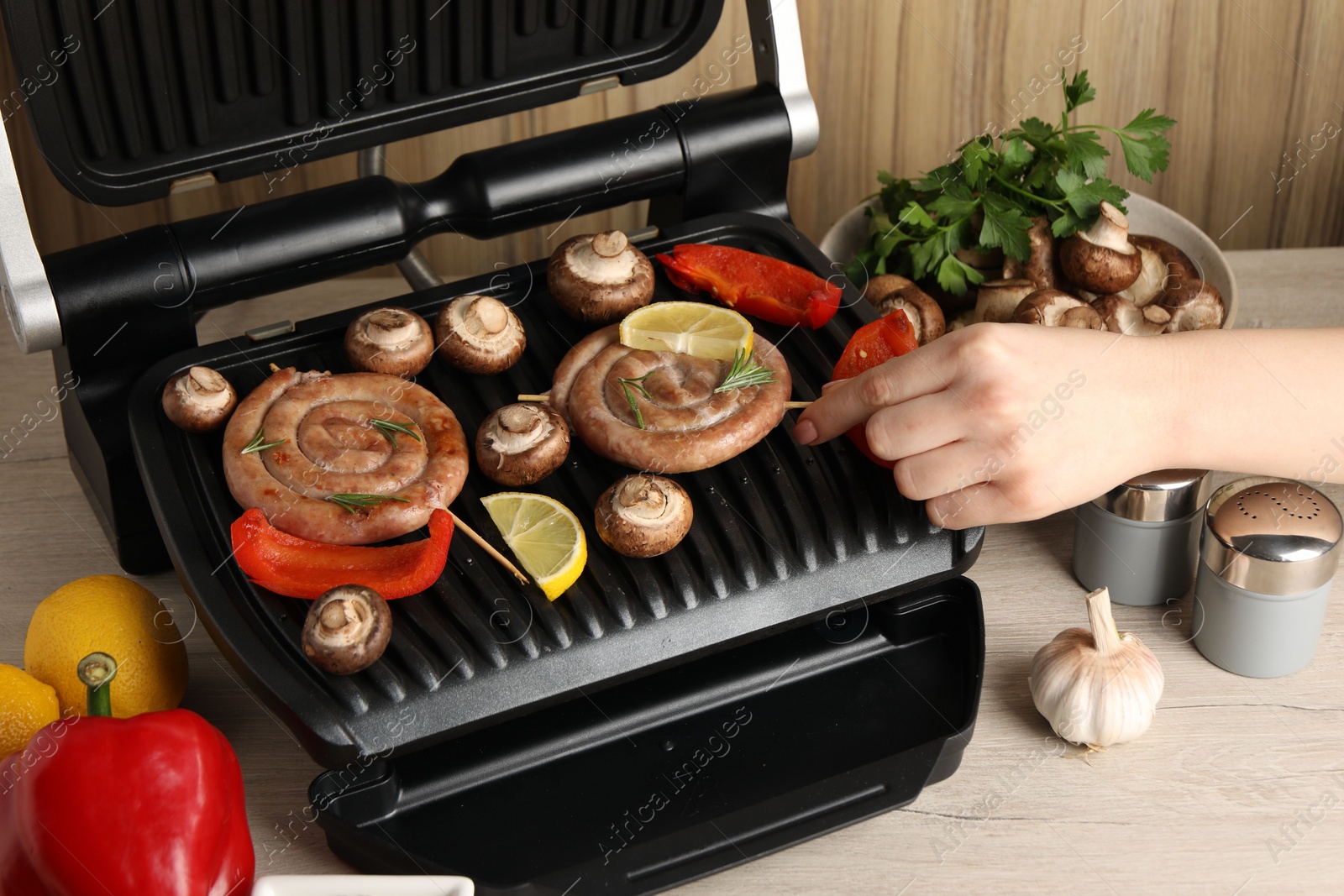 Photo of Woman cooking homemade sausages with mushrooms and bell pepper on electric grill at wooden table, closeup