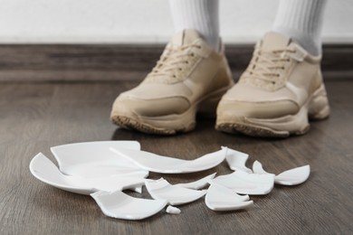 Photo of Woman in sneakers standing near broken plate on floor indoors, closeup