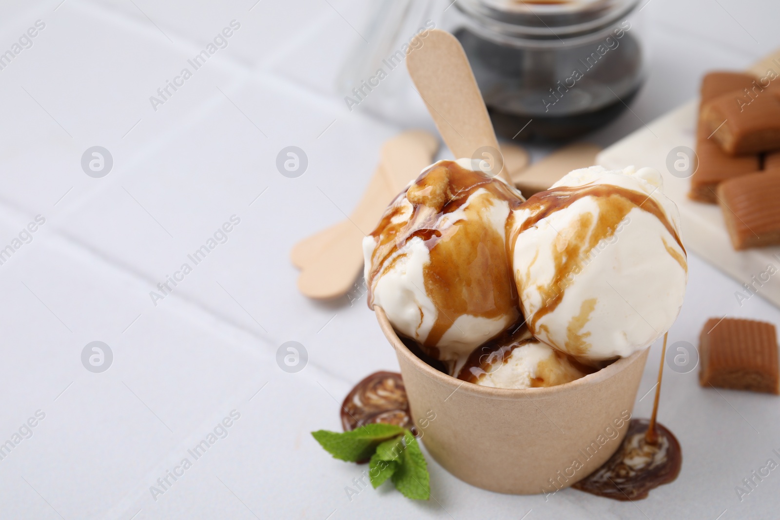 Photo of Scoops of ice cream with caramel sauce in paper cup on white tiled table, closeup. Space for text