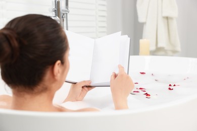 Photo of Woman reading book while taking bath in tub with foam and rose petals indoors, back view