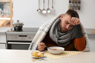 Sick young man eating tasty soup to cure flu at table in kitchen