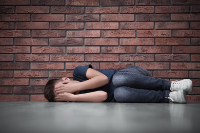 Photo of Sad little boy closing eyes with hands on floor near brick wall. Child in danger