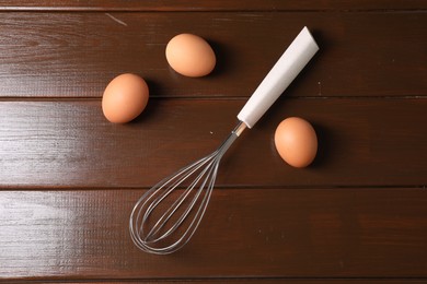 Photo of Metal whisk and raw eggs on wooden table, flat lay