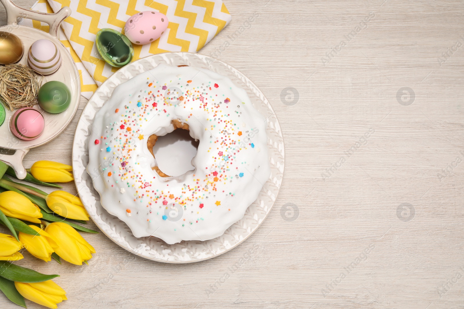 Photo of Easter cake with sprinkles, painted eggs and tulips on white wooden table, flat lay. Space for text