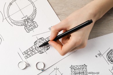 Jeweler drawing sketch of elegant ring on paper at wooden table, top view