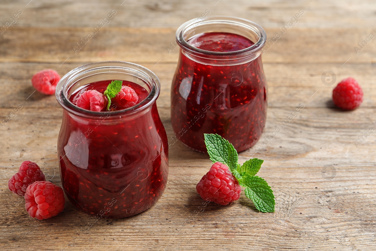 Photo of Glass jars of sweet jam with ripe raspberries and mint on wooden table