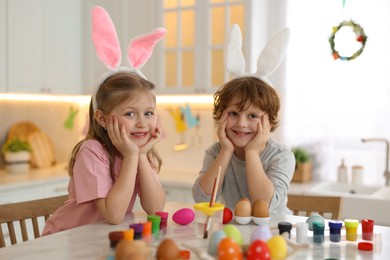 Photo of Painting Easter eggs. Cute children with bunny ears at white marble table in kitchen