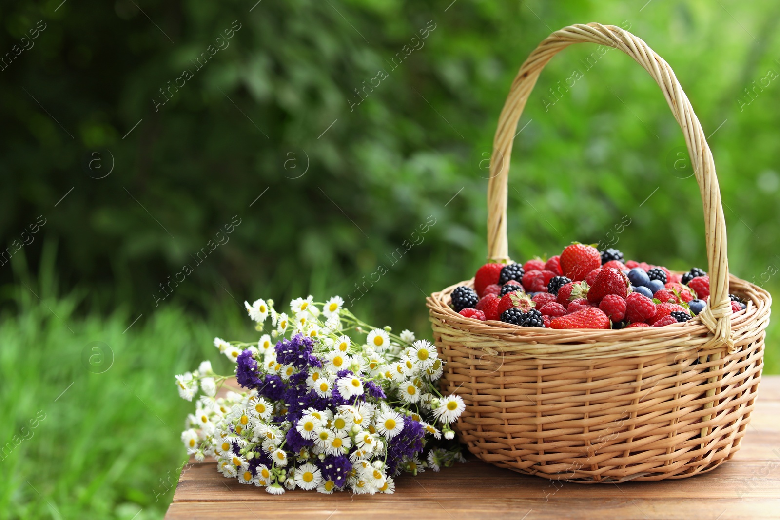 Photo of Wicker basket with different fresh ripe berries and beautiful flowers on wooden table outdoors, space for text