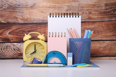 Photo of Different school stationery and alarm clock on white table near wooden wall. Back to school